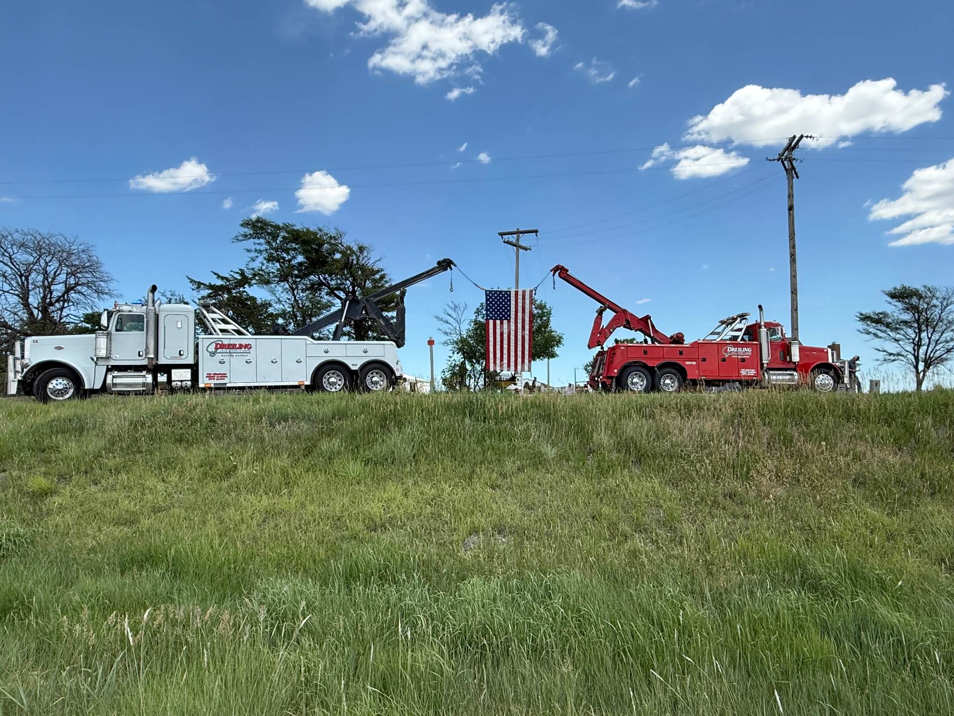 Two Dreiling heavy-duty Peterbilt wreckers, one white and one red, with booms raised holding an American flag between them in a Kansas pasture