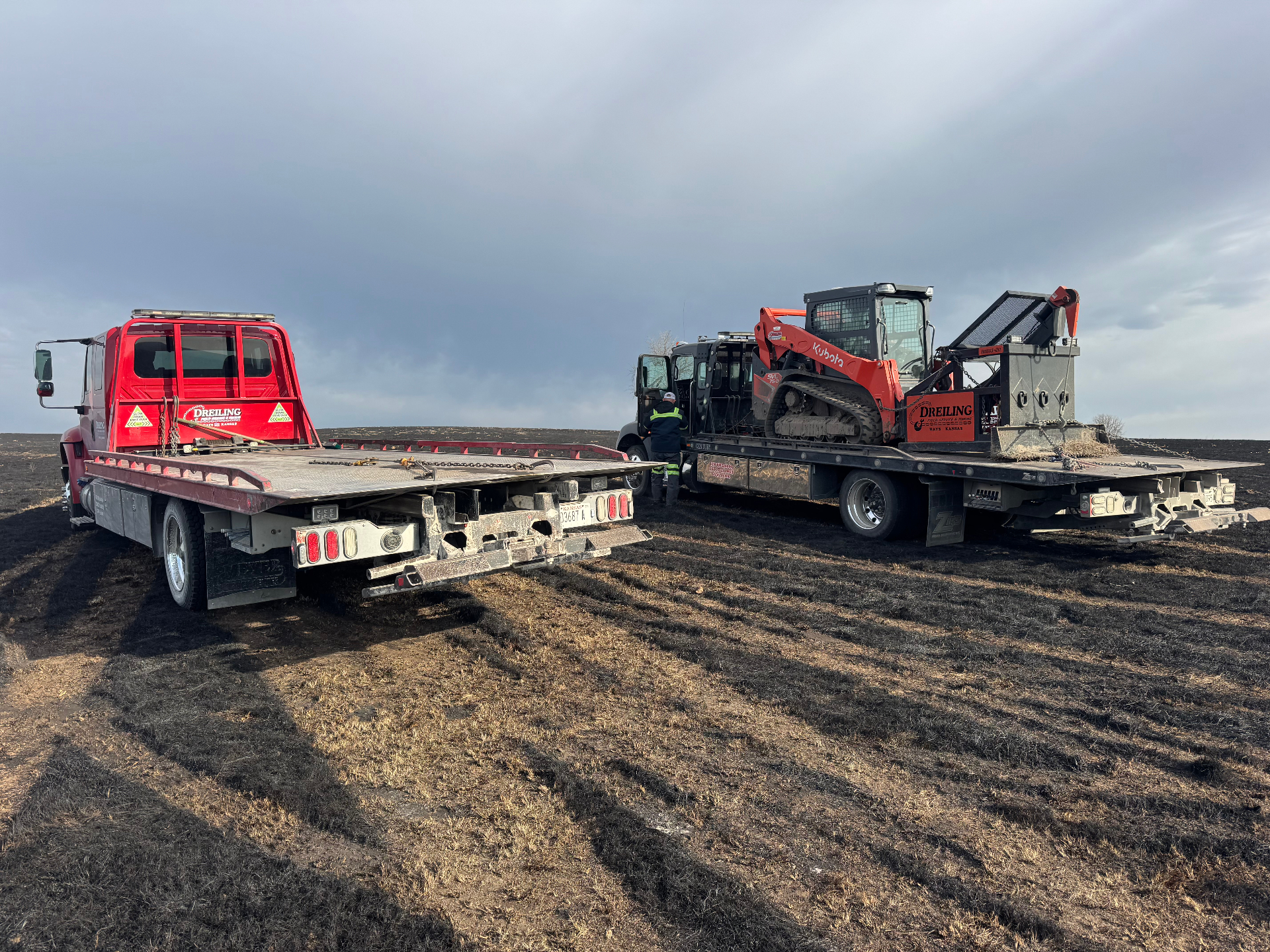 Two Dreiling Field Service & Towing rollback trucks hauling a Kubota skid steer across a burned pasture in Western Kansas