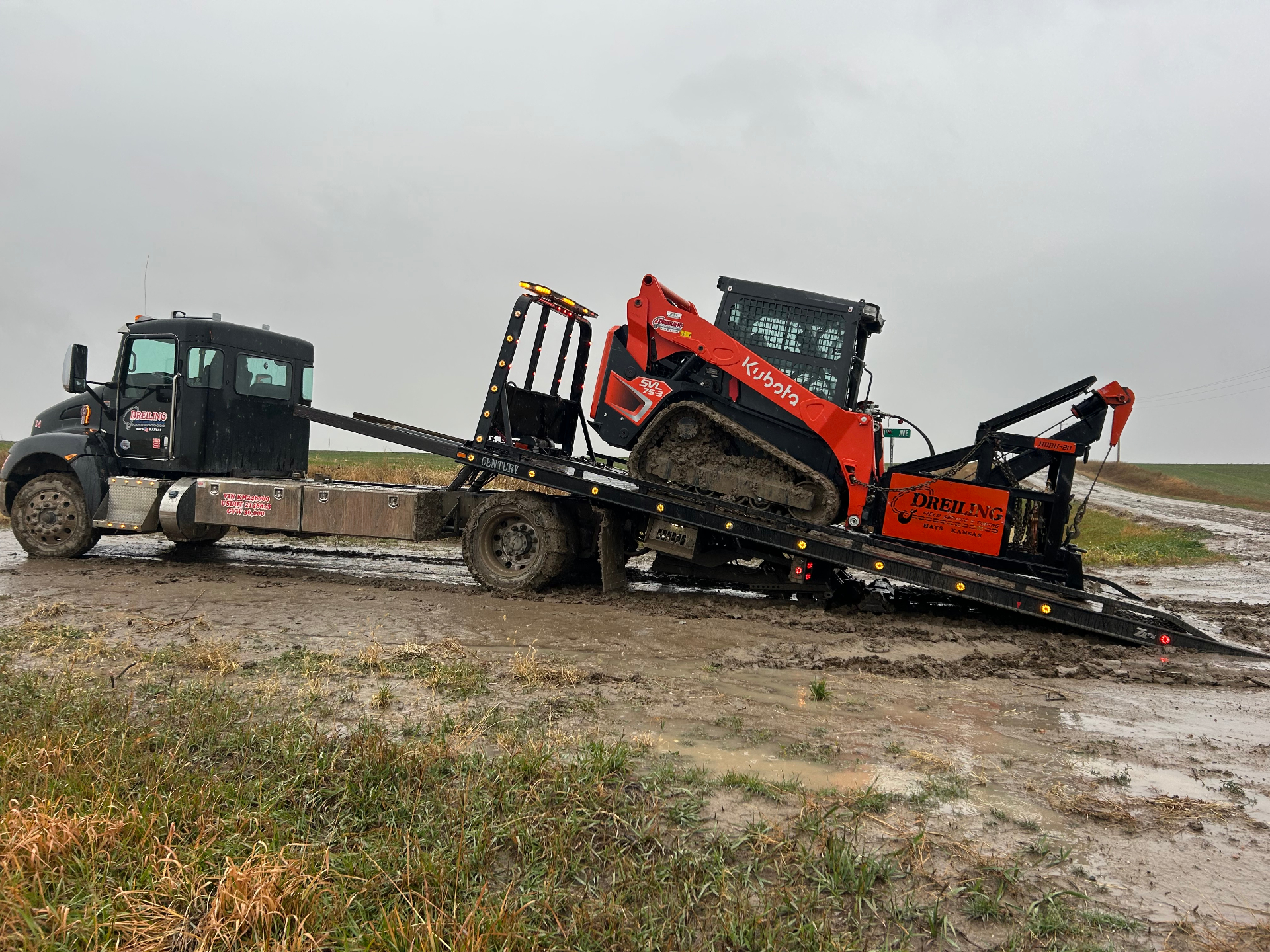 Dreiling Kenworth rollback loading an orange Kubota SVL 75-3 skid steer out of deep mud on a rainy Kansas back road