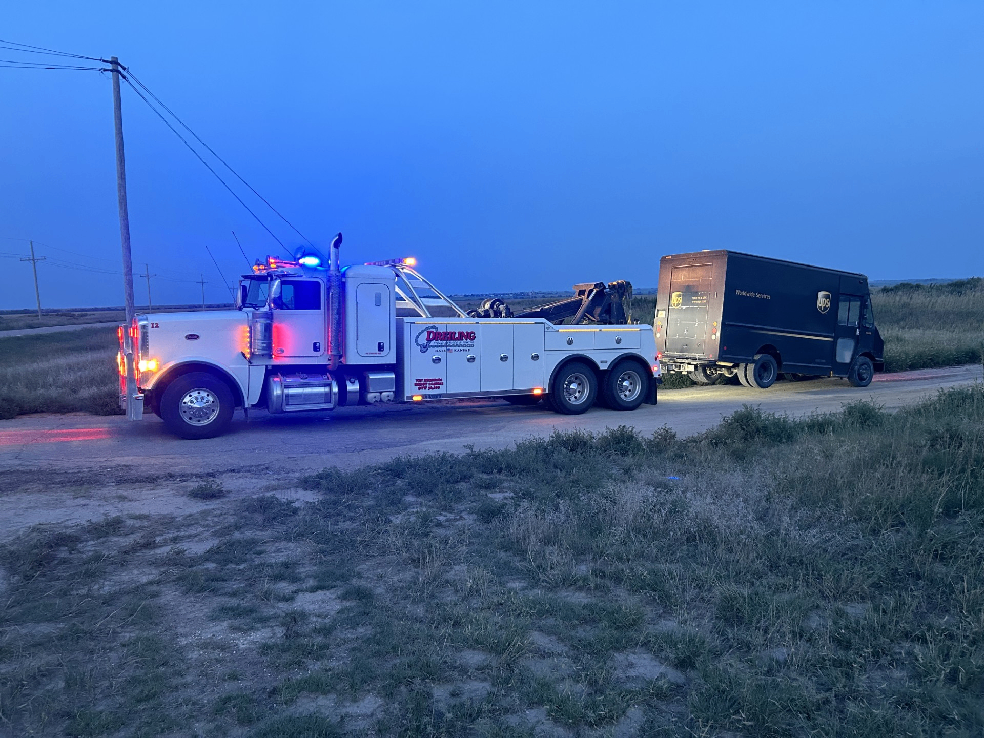 Dreiling Field Service & Towing heavy-duty Peterbilt wrecker towing a UPS delivery truck at dusk near Hays, Kansas